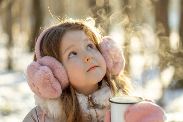 在一个阳光明媚的冬日早晨，一个戴着粉色毛皮耳机，一头金色长发的小女孩站在森林里，手里拿着一杯热茶，看着茶里冒出来的蒸汽图片下载