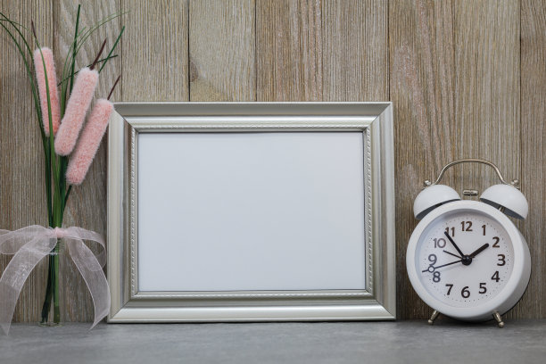 银色框架模型与白色闹钟和花瓶中的花对艺术或复制空间的木墙。图片下载