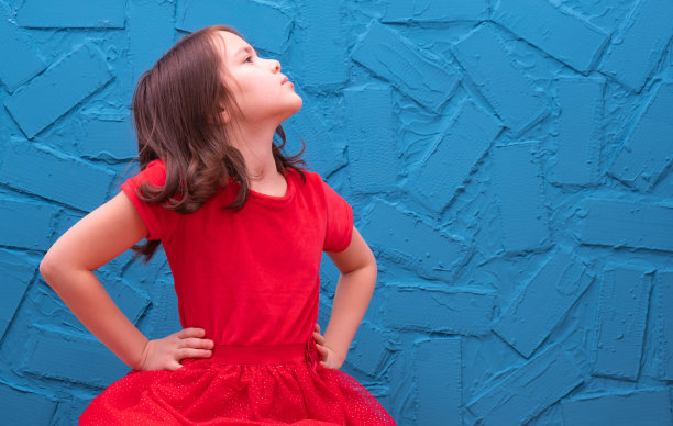 一个穿着红色连衣裙的小女孩靠墙坐着，她傲慢地坐在蓝色的背景上图片下载