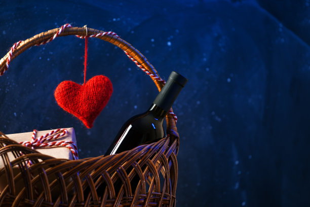 情人节节日贺卡。一瓶红酒，一个柳条篮子里的礼盒，一颗象征爱情的红色针织心。浪漫的晚餐图片下载