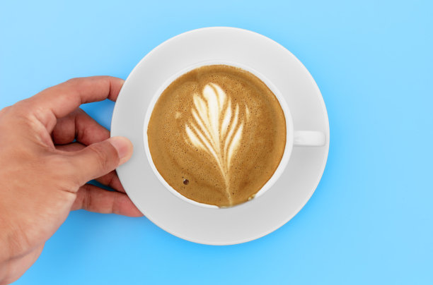 俯视图手握白色咖啡杯与卡布奇诺咖啡和奶油形状在白色杯子。在咖啡店的蓝色桌子上，手持咖啡杯的男人。图片下载