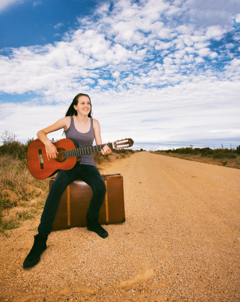可爱的年轻女子弹吉他旁边的土路，她坐在她的手提箱在农村的设置图片下载