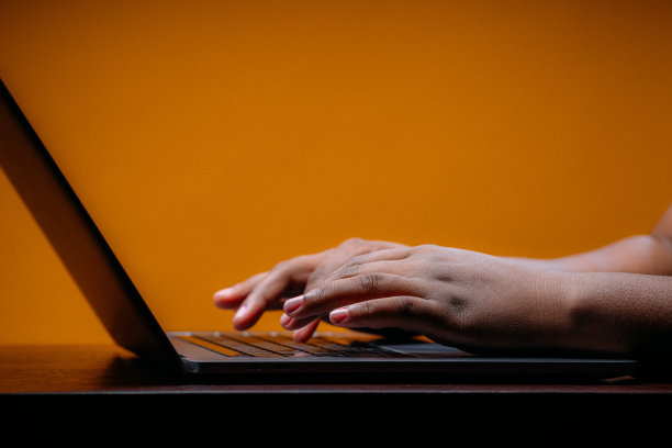 年轻的非裔美国职业妇女的手打字笔记本电脑在戏剧性的灯光下，在一个黄芥末色的工作室背景与复制空间图片下载