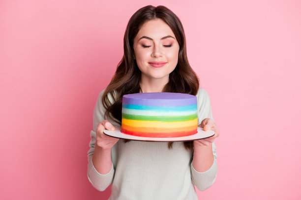 迷人的波浪发型女士的照片，手臂抱着蛋糕，闭着眼睛闻，在淡粉色的背景上孤立图片下载