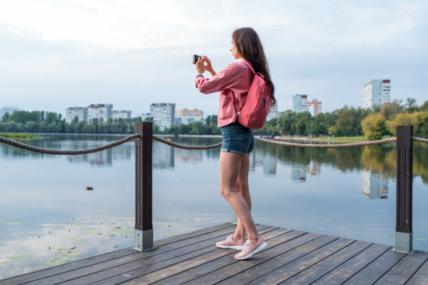夏天，女孩在城市公园附近的水库、池塘、河流和湖泊，用智能手机录制视频照片。观光和旅游。风景摄影。牛仔短裤夹克背包。图片下载