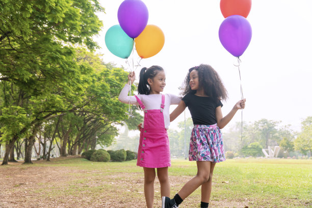 两个欢快的小女孩拿着彩色气球，一起站在绿色的草地上。可爱可爱的女孩玩气球，感觉自由，而看着对方在公园里。友谊的概念。图片下载