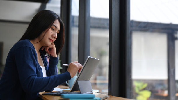 年轻女性自由设计师坐在靠窗的工作台上，在平板电脑上工作。图片下载