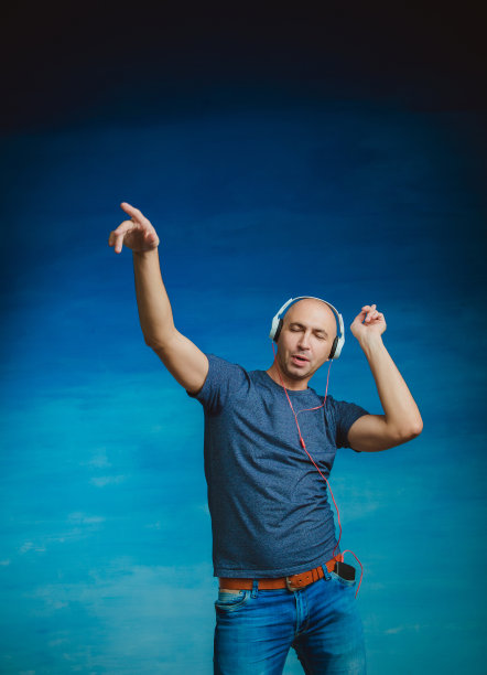 一个秃顶的男人戴着大耳机，闭着眼睛在蓝色背景上听音乐。闭着眼睛跳舞，用手打手势图片下载