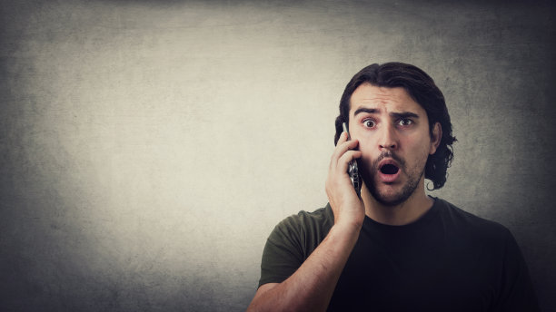 震惊的年轻人，在一个手机通话，瞪着大眼睛和嘴巴孤立在灰色的墙壁背景，与拷贝空间。一个惊讶的家伙在电话里听到了坏消息图片下载