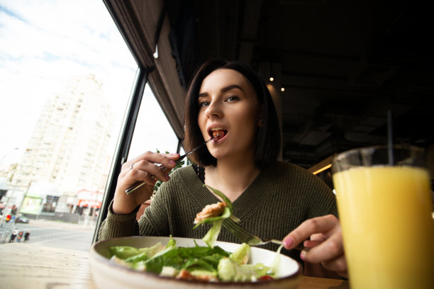 深色头发的女人愉快地吃着凯撒沙拉。前面有一杯模糊的鲜橙汁。深色的背景和明亮的大窗户。商务午餐时间概念。美味和健康的食物。图片下载