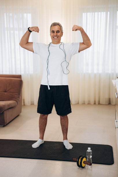 骄傲的成熟男子展示他的肌肉。高强度训练的概念。健康的长者显示二头肌和微笑。照亮客厅的背景。英俊的男人经常锻炼以获得健康的身体。完整的长度。图片下载