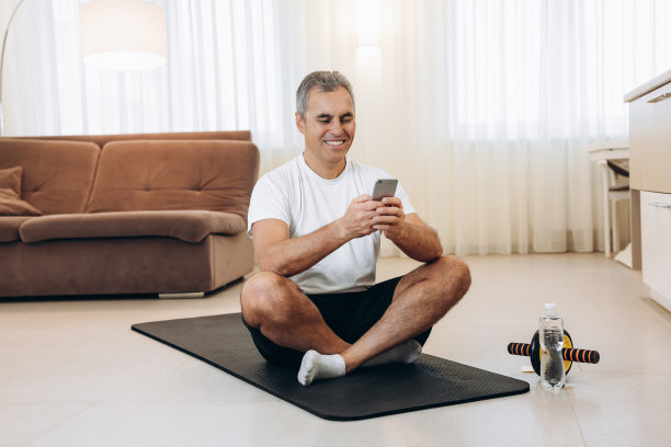懒惰的老人在智能手机上阅读表情包时大笑。放下你的手机，如果你想要一个强壮健康的身体!拖延在健身。老男人坐在瑜伽垫上，用手机发短信。图片下载