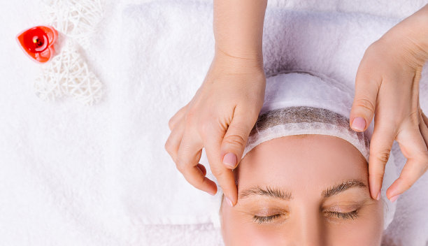 一张放大的照片，一个女人闭着眼睛，正在做脸部和头部按摩。白色毛巾和心形香薰蜡烛。图片下载