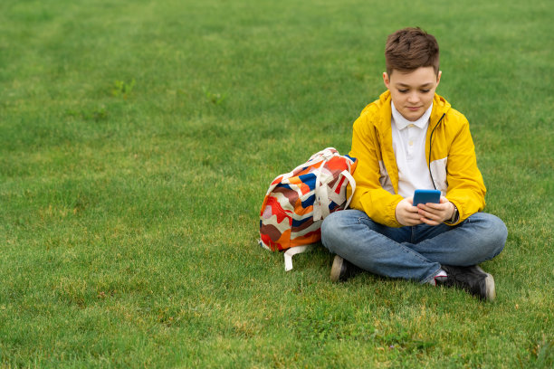 一个男生坐在草坪上，手里拿着智能手机。穿着黄色夹克的现代少年图片下载