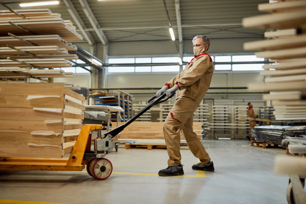 在木工车间，带面罩的手工工人正在拉动带木板的托盘千斤顶。图片下载