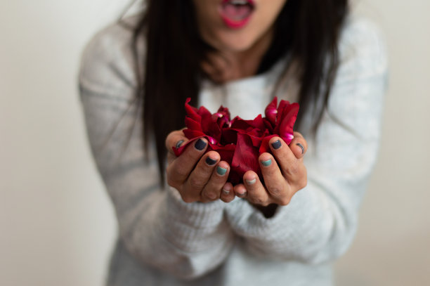 一个女人拿着一朵红玫瑰的花瓣，用手吹着。选择性地聚焦在花瓣上。爱的概念图片下载