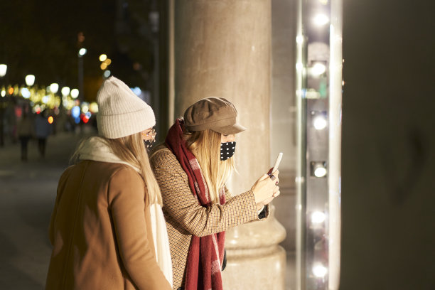 一个晚上，在一座城市里，两个年轻的妇女，穿着冬装，看着一家商店的橱窗。其中一个用手机拍了张照片。图片下载