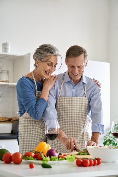 快乐的60多岁的素食夫妇穿着围裙在家准备健康的饮食沙拉。微笑成熟的高级家庭拥抱，一起做饭，一起切新鲜的有机蔬菜在厨房。图片下载