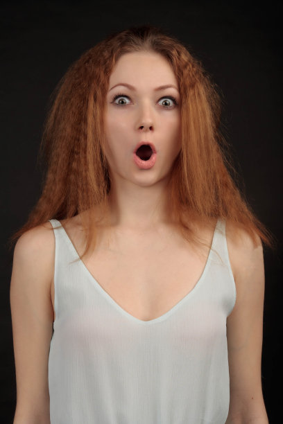 可爱的年轻女子穿着白色连衣裙看起来惊讶地看着镜头，她的嘴张着。情感工作室肖像对黑色的背景。图片下载
