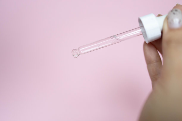 一个美丽女人的双手将血清胶原蛋白保湿液滴入清澈的瓶子中，形成柔和的粉色背景。护肤护理面部精油，维他命c，美容spa图片下载