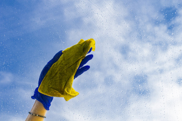 在一片模糊的蓝天和云朵的背景下擦洗窗户玻璃，玻璃上有水滴，一个女人拿着抹布的手。春天的概念，纯洁和更新图片下载