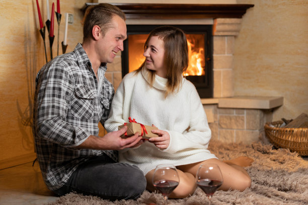 男孩给了他的女朋友一个节日礼物，他们坐在壁炉旁边的地板上，看着对方的眼睛。在一个舒适的家里庆祝情人节的概念。图片下载