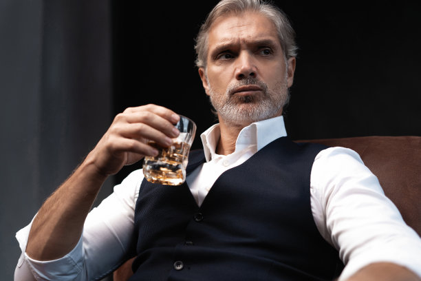 在黑暗的房间里，帅气优雅的成熟男子拿着一杯威士忌的肖像。图片下载