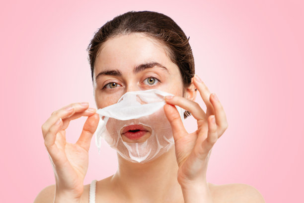 一个年轻美丽的女人的肖像从她的脸上摘下面具。粉红色的背景。保湿、美容、美容的概念图片下载