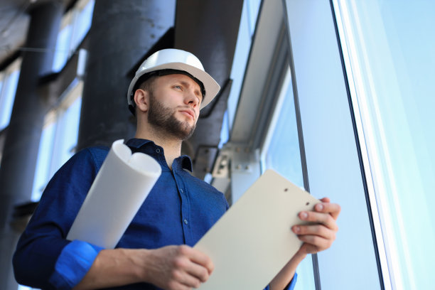 自信的年轻商人戴着安全帽，站在室内检查蓝图。图片下载