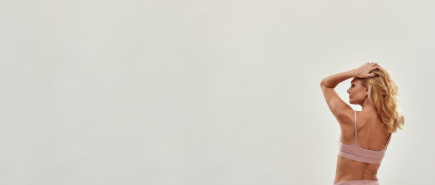 一个半裸美丽的中年金发女人在内衣站在工作室的灰色背景和看别处的后视图图片下载