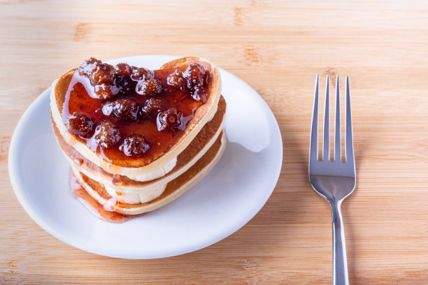 白色的盘子上放着自制的带浆果酱的心型煎饼，木制的背景上放着一把叉子图片下载