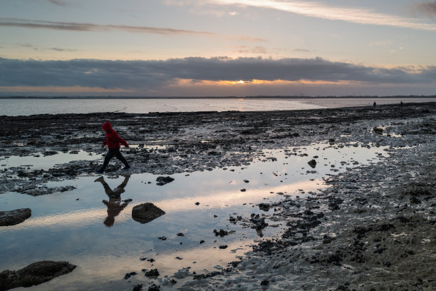 夕阳西下时海滩上的一个岩石池，映照着天空。一个男孩带着水中的倒影跑了过去图片下载