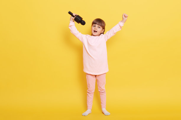 小女孩拿着麦克风在黄色背景下唱歌，一直举着手，安排音乐会，为某人表演，在卡拉ok唱歌，扎着辫子的孩子穿着随意。图片下载