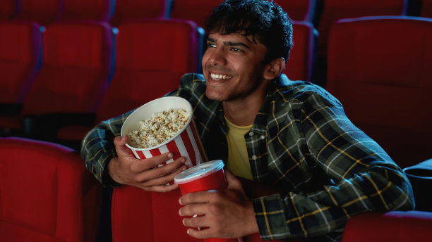 一个快乐的年轻人的肖像，微笑着，拿着饮料和爆米花篮子，而独自看电影在空的剧院礼堂图片下载