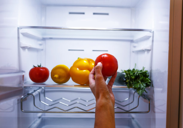 一个女人的手从冰箱的架子上拿起一个西红柿。冰箱里装满了蔬菜和水果。健康食品。健康的生活方式的概念图片下载