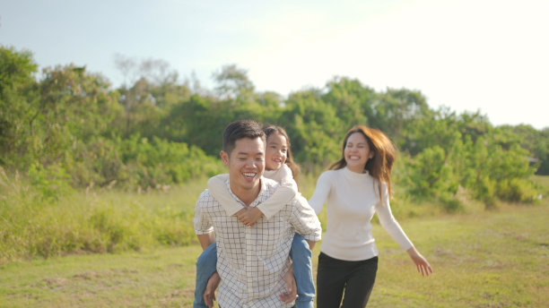快乐年轻的亚洲父亲背着他的小女孩，而母亲跑和她在公园玩，在绿色的花园一起享受我们自己。一家人在大自然中享受阳光明媚的秋日。图片下载