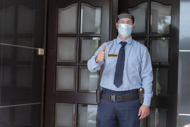 一名在安全建筑工作的男子穿着制服，戴着生物安全措施用面具保护自己，看着一幅肖像中的摄像机图片下载