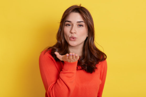 漂亮迷人可爱的女性的特写，风骚开朗的女孩噘嘴送温柔的空气之吻在黄色的背景，看着相机。图片下载
