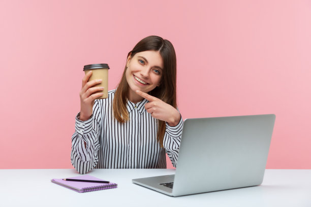 微笑充满活力的女办公室职员指着纸杯和饮料坐在工作的笔记本电脑，喝咖啡来激励图片下载