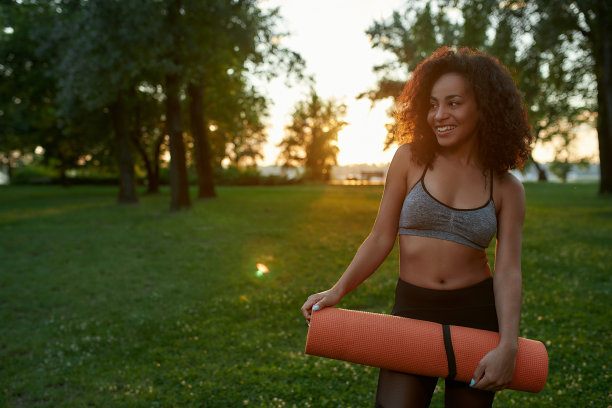 在户外体能训练。年轻快乐的混合种族妇女穿着运动服拿着瑜伽垫，微笑着看一边，而站在公园在清晨图片下载