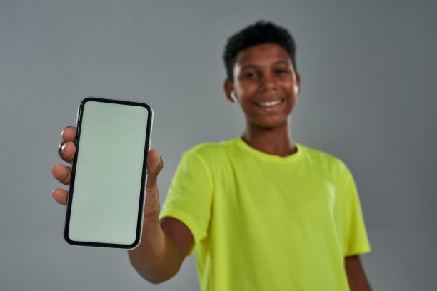 一名微笑的非洲少年身穿霓光灯t恤，灰色背景下站着，屏幕上显示着智能手机图片下载