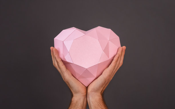 多边形心形纸制成。灰色背景上一个男人手中的粉色心脏图片下载