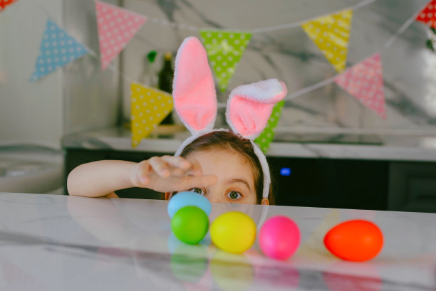 有趣的兔耳朵小女孩坐在厨房的桌子旁玩彩色的复活节彩蛋。假期的概念。图片下载