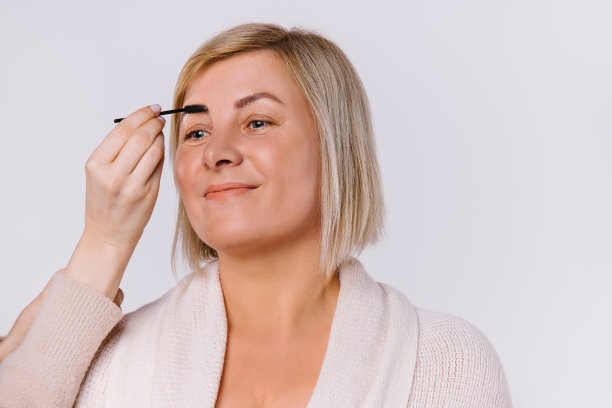 白色背景上画着温柔的妆容，面带微笑的老妇人。这位大师的手用化妆刷画眉毛。图片下载