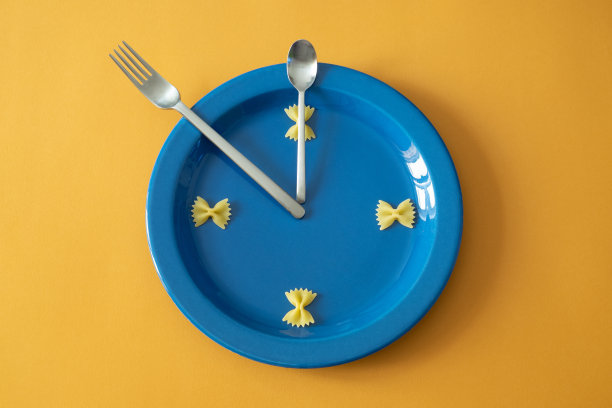 由一个蓝色的圆形盘子、两个餐具和生的意大利面组成的手表，以表示时间，表达了午餐的概念。图片下载