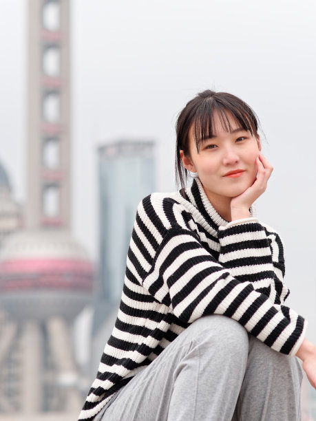 冬日里，一名中国年轻女子手捂着脸对着镜头微笑，背景是上海的地标建筑。图片下载