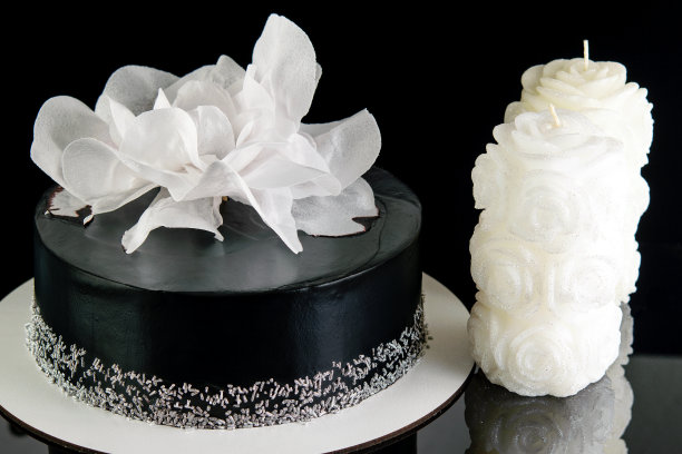 美味的甜点。自制的节日黑色蛋糕，装饰着白色的大花，在黑色的反光背景上插着白色的蜡烛。图片下载