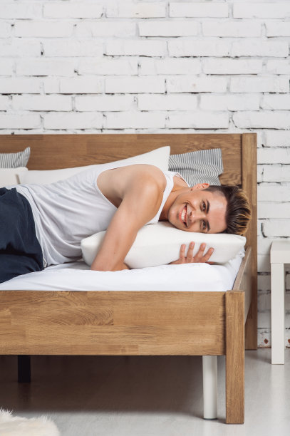 一个微笑的男人躺在白色的枕头和木床上的床垫上。舒适睡眠的概念。骨科床垫和枕头图片下载