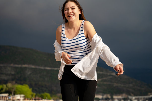 快乐的年轻女子身穿白衬衫和条纹上衣，在度假小镇周围散步，笑着对着山脉和黑色的天空摆姿势。暴风雨前的平静图片下载