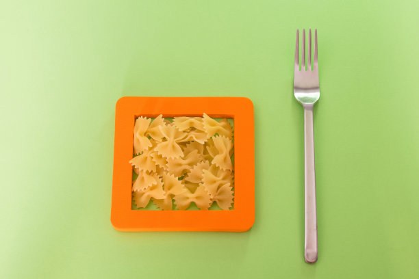 广告或厨房陈设的创意背景，一堆生蝴蝶结形状的短面团放在一个方形的橙色框架里，右边放着一个钢叉子。图片下载